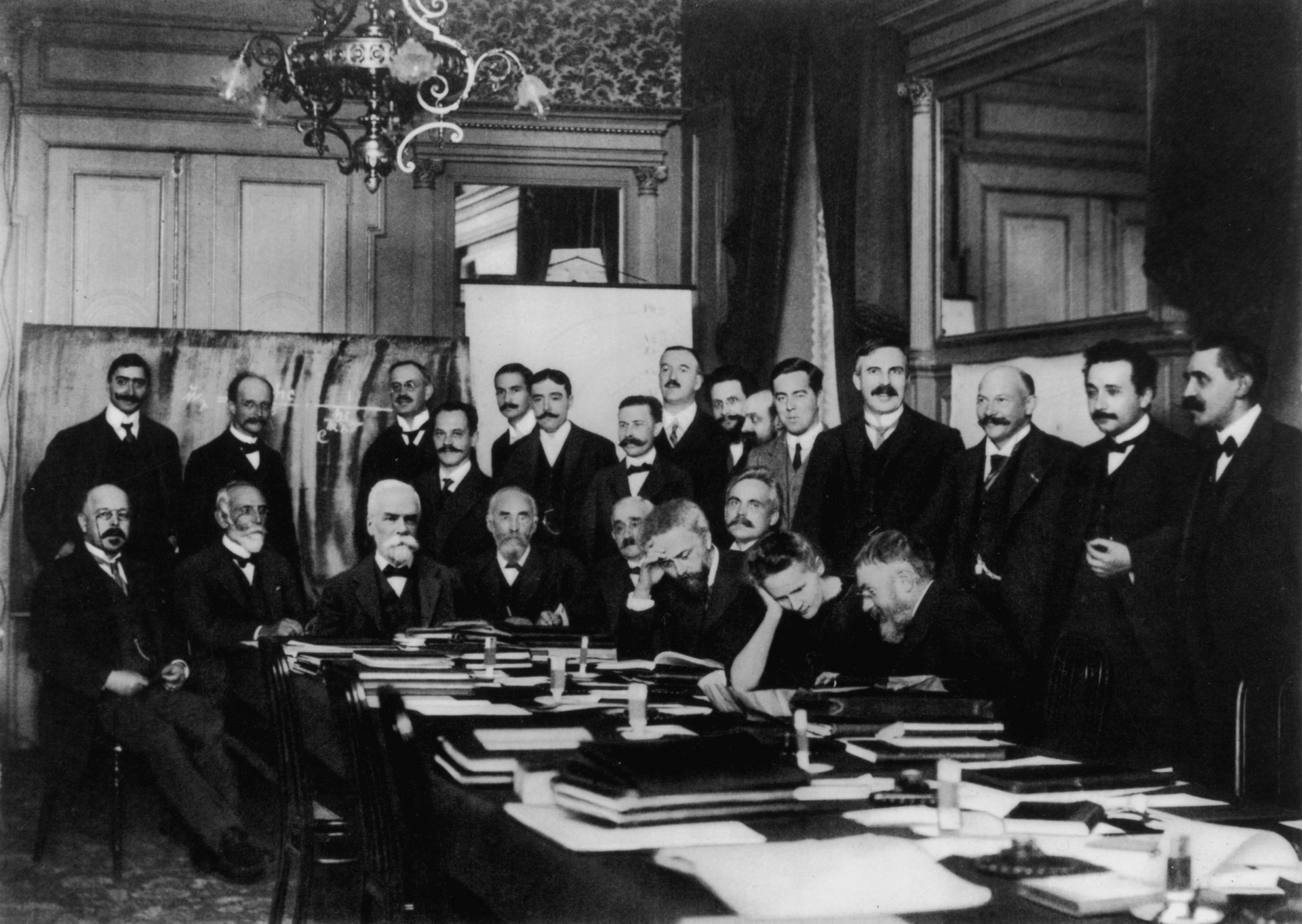 This is a photograph of a meeting of 23 men and Marie Curie at the 1911 Solvay Conference. They all sit or stand around a table covered in notebooks and other work materials.