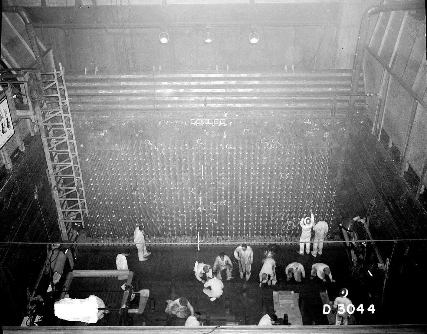 This photograph shows men in white around a giant reactor making adjustments.