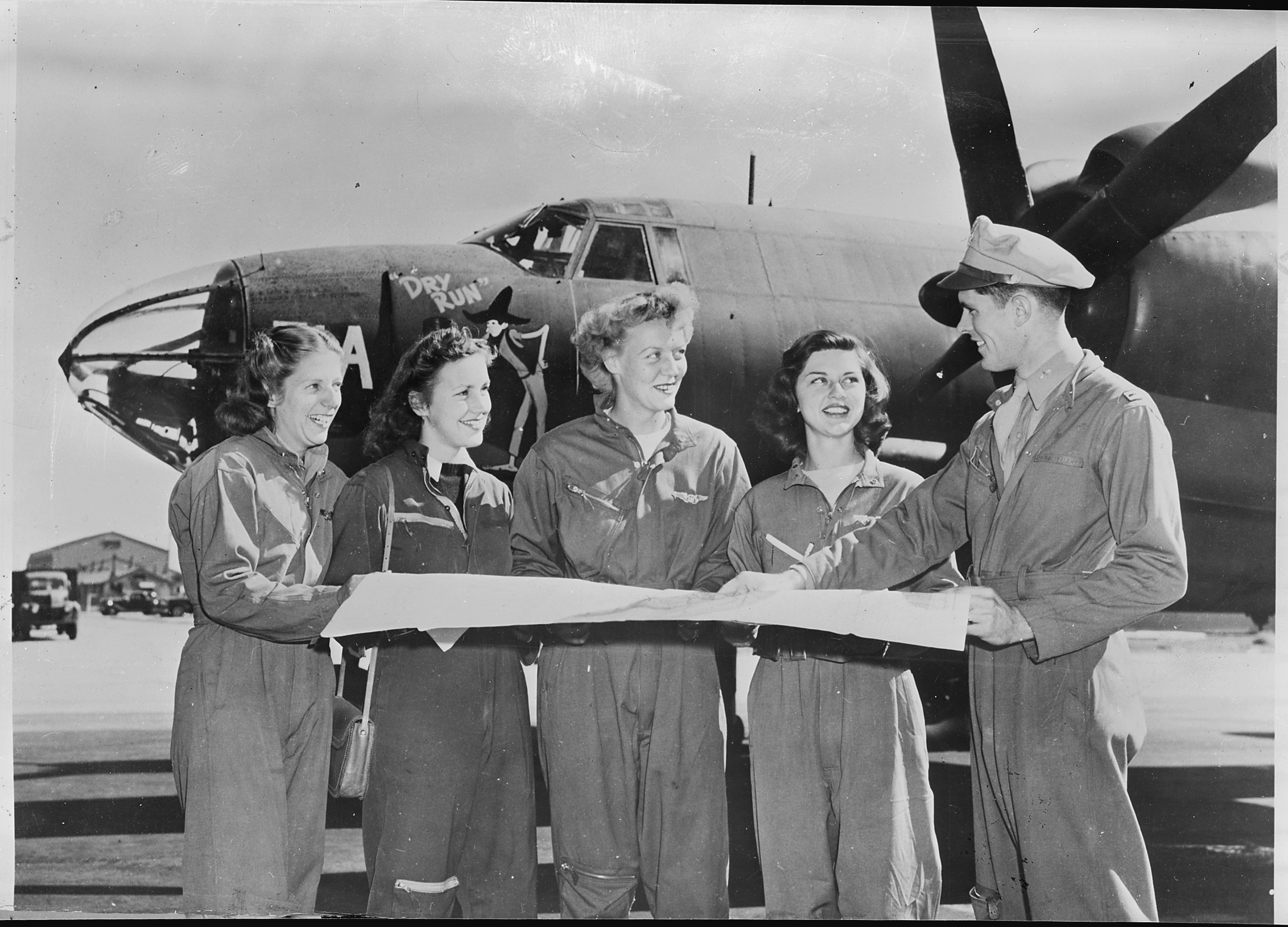 Photograph of four members of the United States Women's Airforce Service Pilots (WASPs) receive final instructions as they chart a cross-country course on the flight line of U.S. airport. 