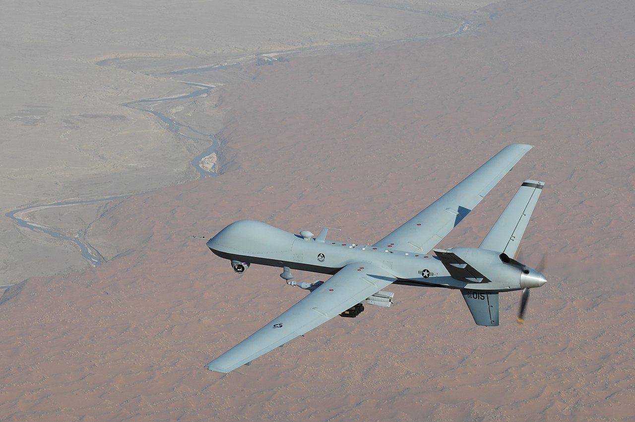 Color photograph of a grey drone flying in air, with the rugged land of Afghanistan visible underneath.