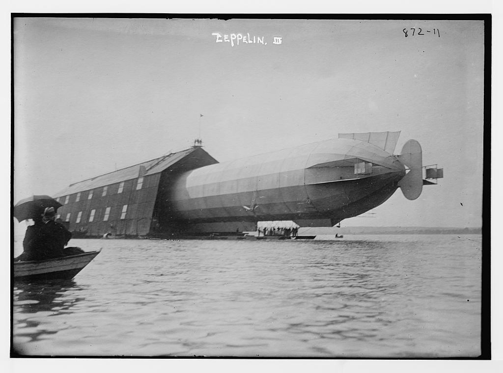 Photograph showing water, with shed floting and a large dirigible sticking out of the shed. Labelled "zeppelin"; there is another person visible in foreground on boat with umbrella.