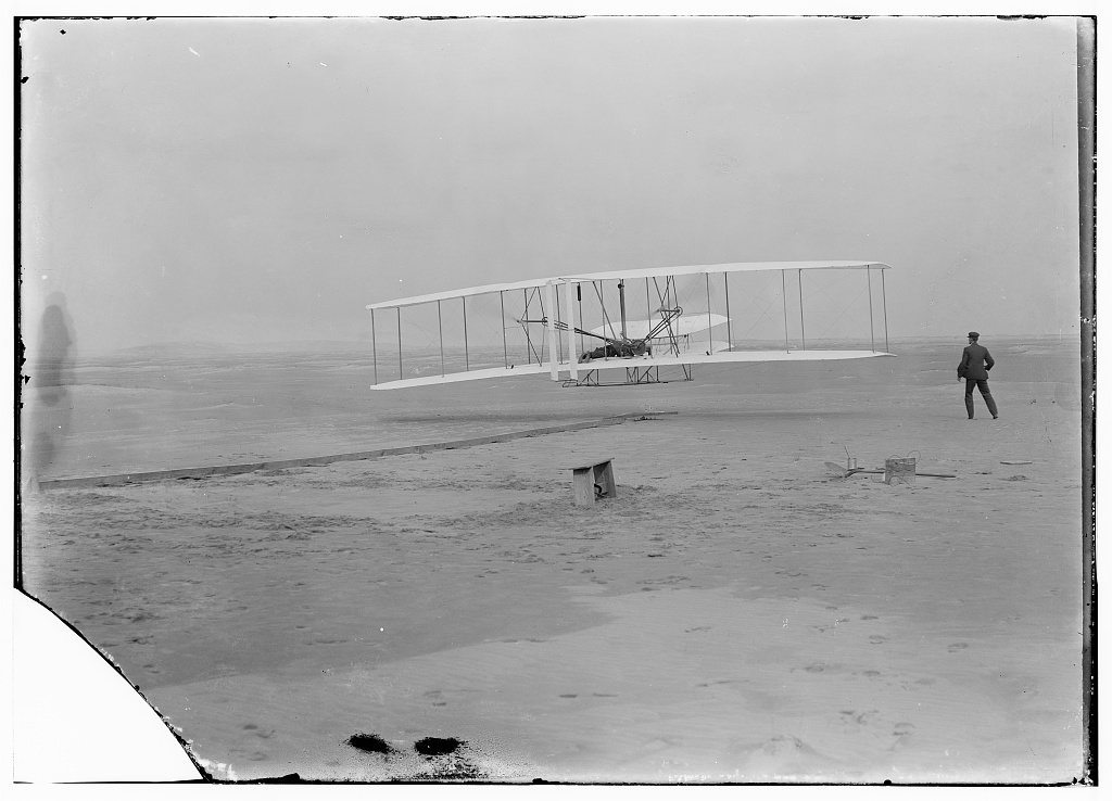 First flight Black and white photograph showing airplane just barely off the ground on beach as man stands alongside.