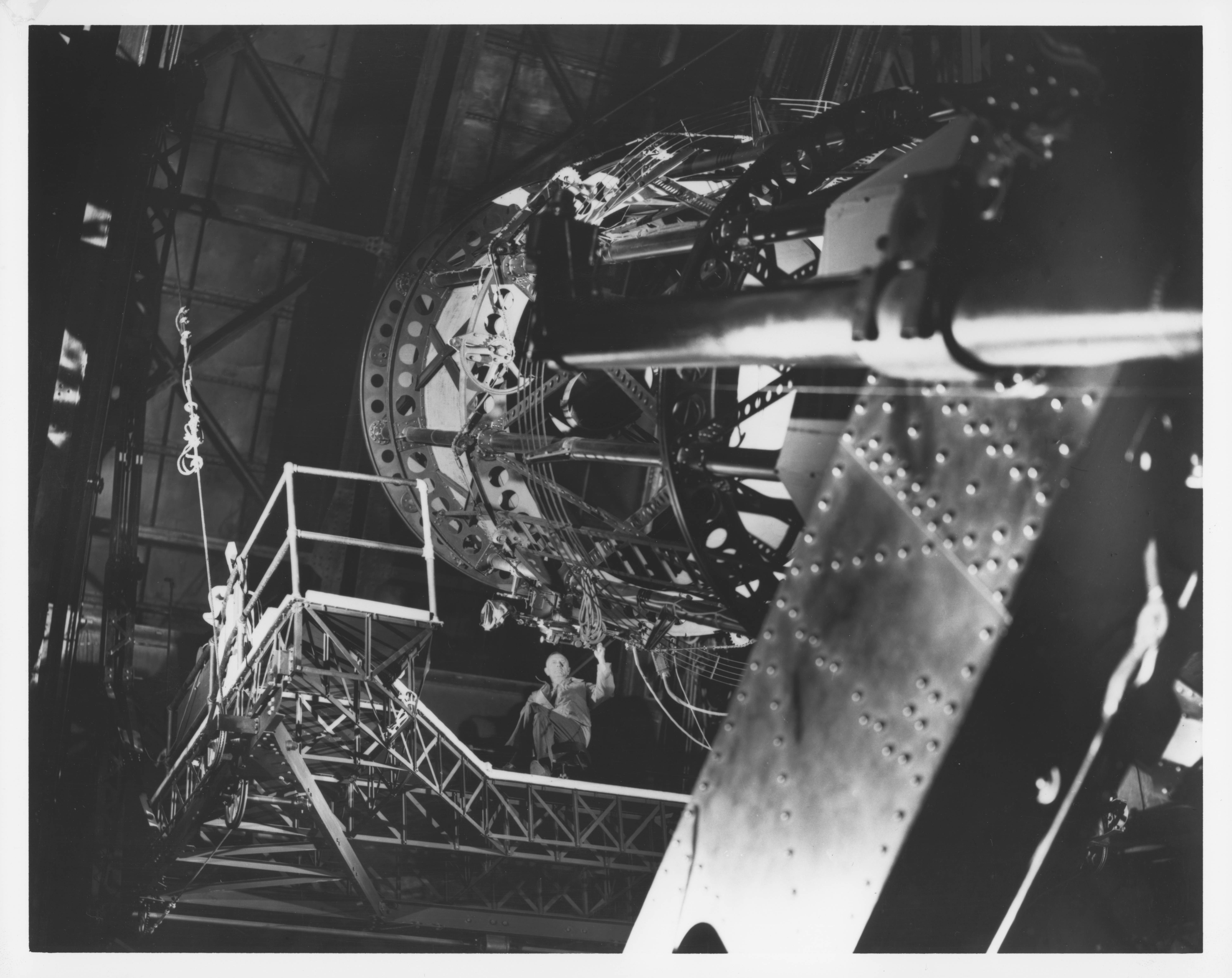 A man in the center is seated on a platform with his hand on a giant telescope that takes up the majority of the image, shot from below looking up.