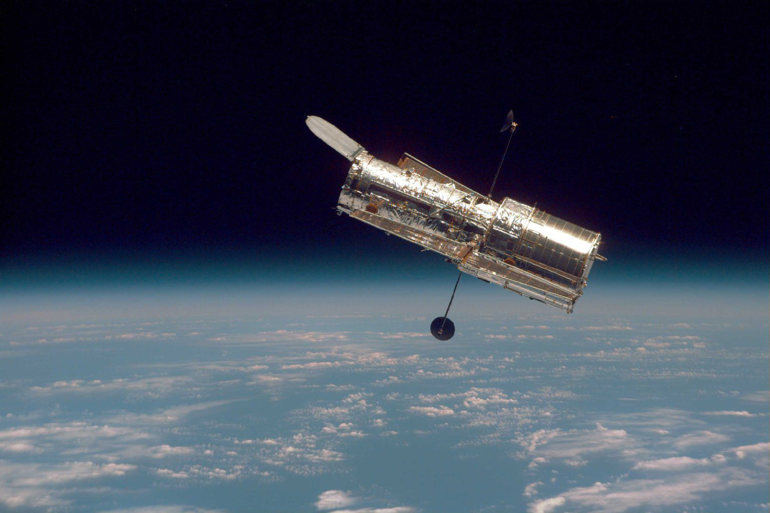 Hubble satellite in outer space, showing its shiny exterior against the backdrop of Earth's atmosphere below and dark space above. 